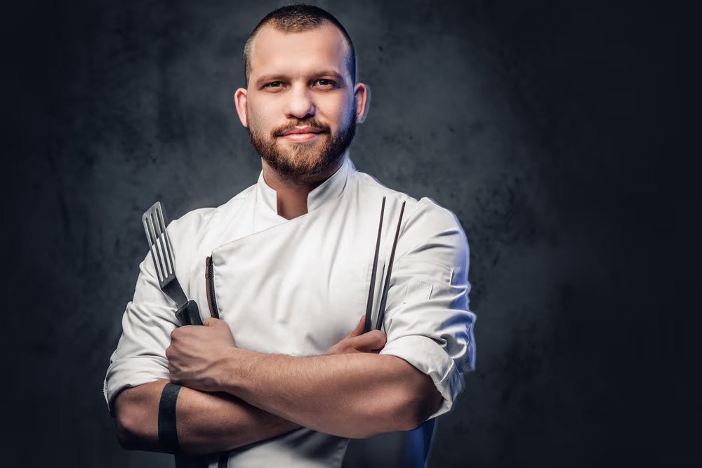 Chef Daniel Reyes in a white chef's jacket, arms crossed, holding kitchen tools